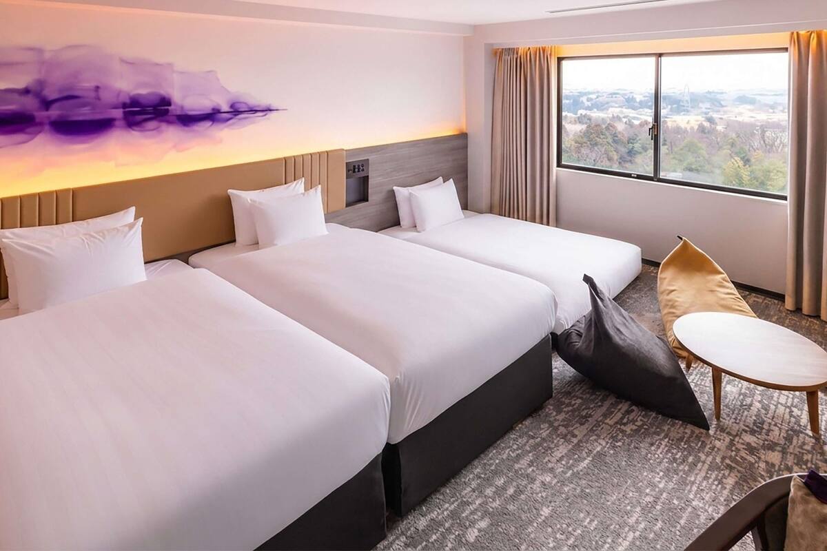 A hotel room with two beds, a small table, a chair, a large window with a view, and a modern wall art piece.