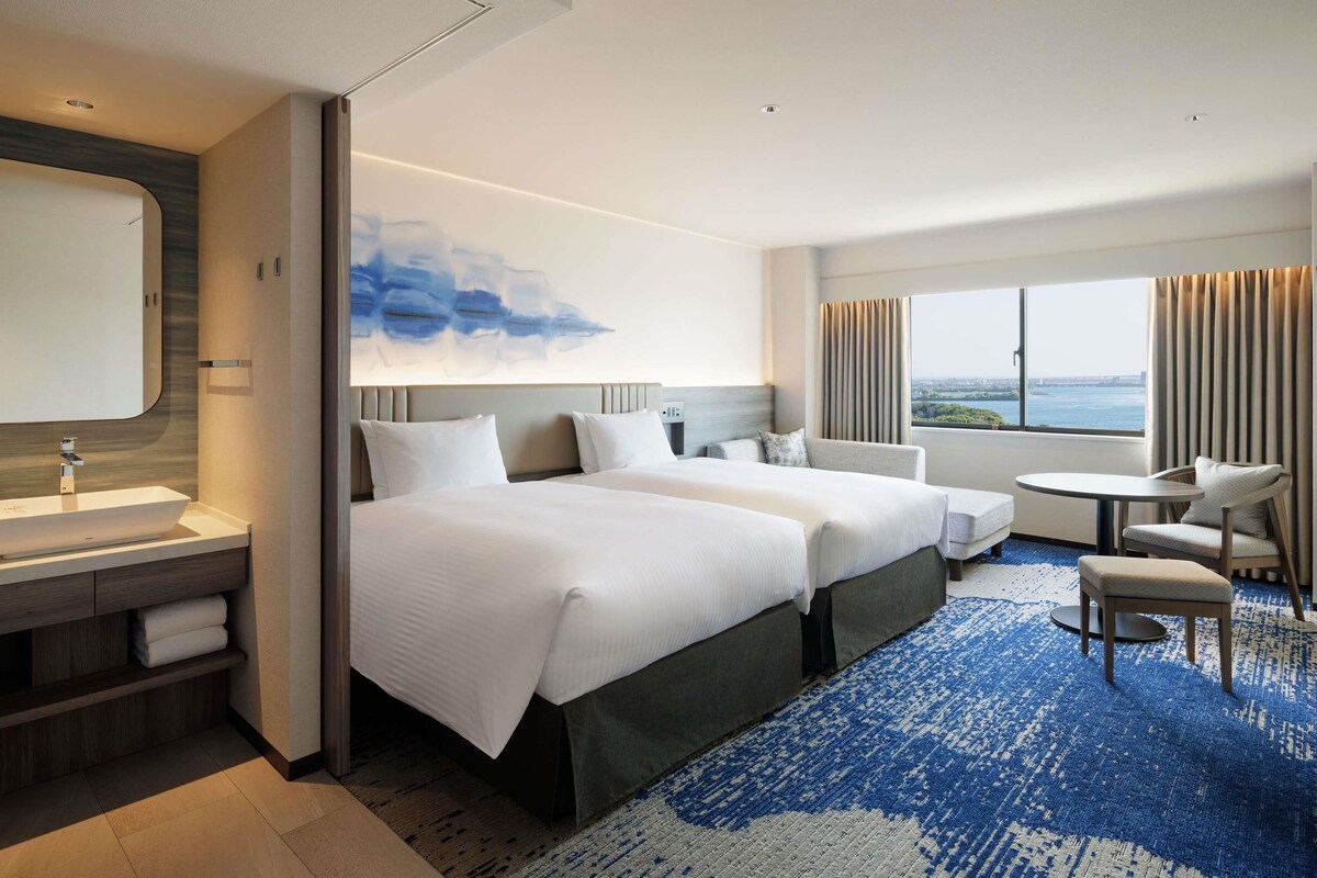A hotel room with a large bed, a desk, a chair, and a view of the city.