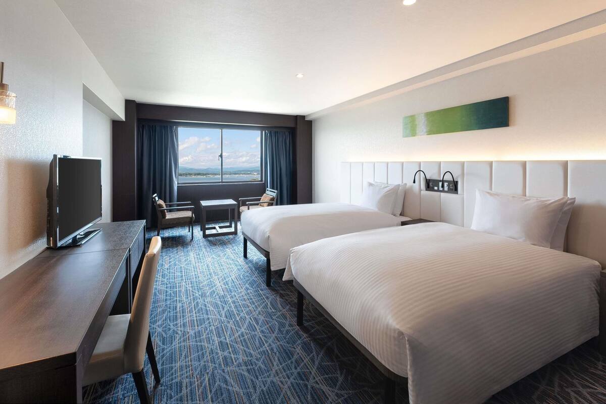 A hotel room with two beds, a desk, a TV, and a large window with a view.