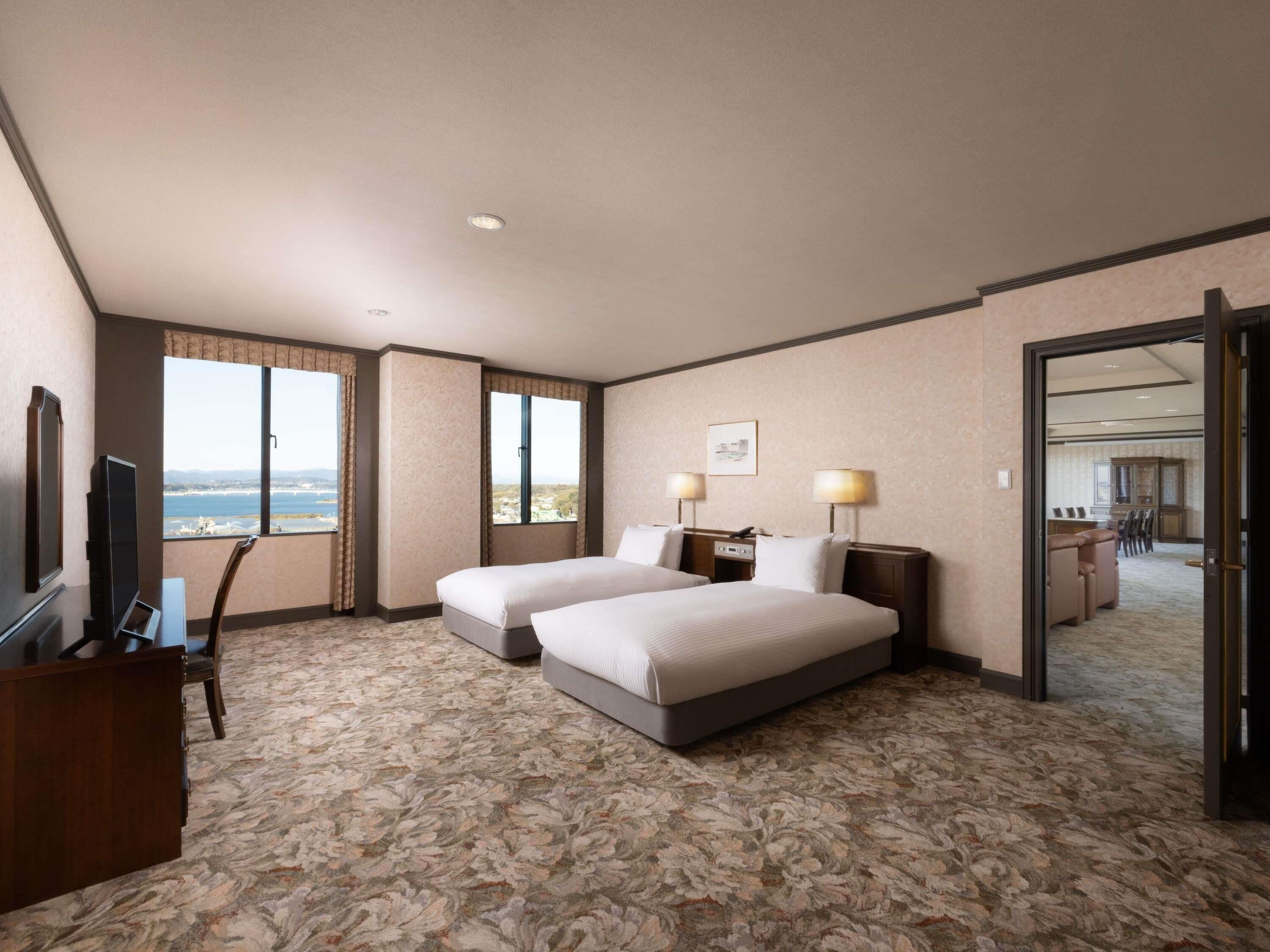Ein Hotelzimmer mit zwei Betten, einem Schreibtisch mit Stuhl, einem Fernseher und Blick auf Stadt und Meer.