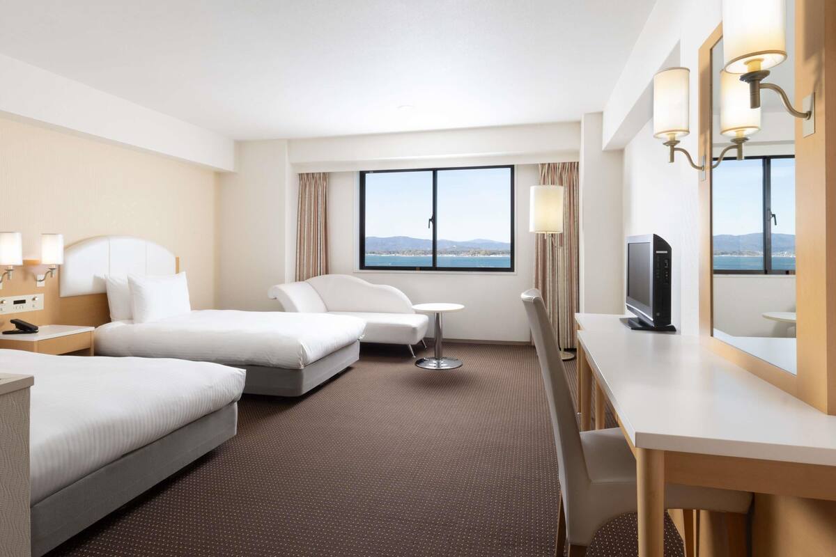 A hotel room with two beds, a desk, a chair, a TV, and a large window with a view.