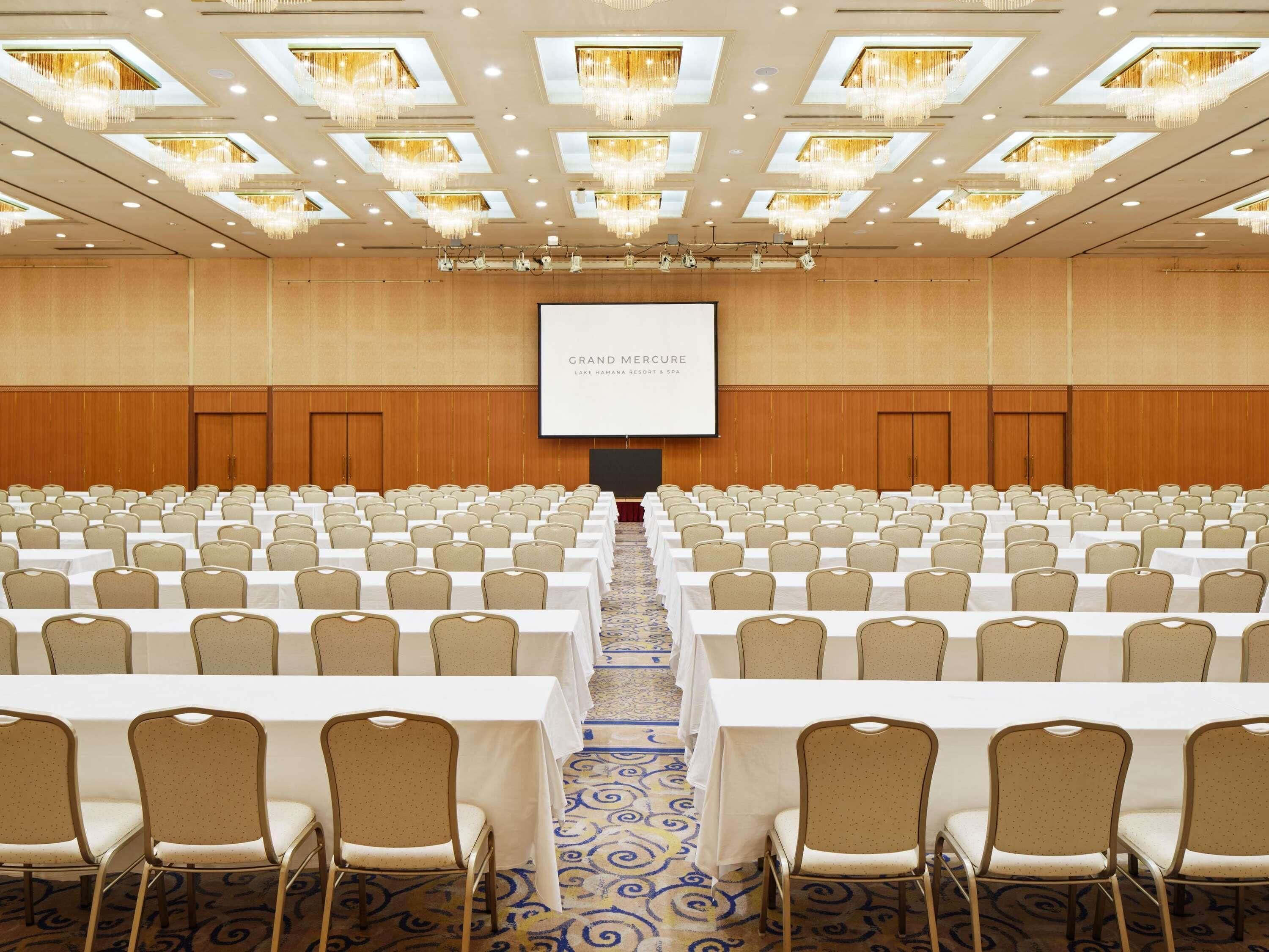 A conference room with rows of tables and chairs, a large screen, and chandeliers.