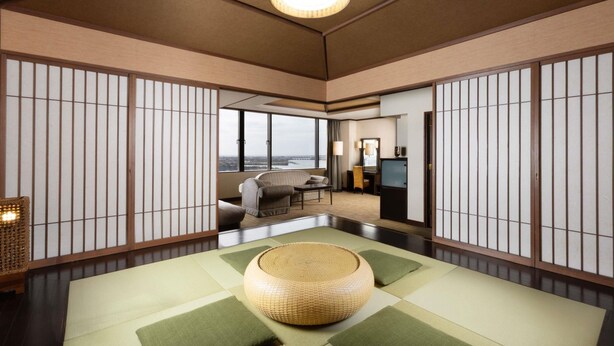 Quarto japonês tradicional com tatames, um banquinho de vime e vista para a cidade através de grandes janelas.