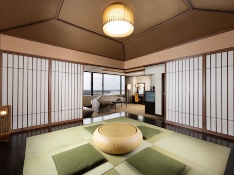 A traditional Japanese-style room with tatami flooring, a woven rattan ottoman, and a view of a cityscape through large windows.. Grand Mercure Lake Hamana Resort & Spa