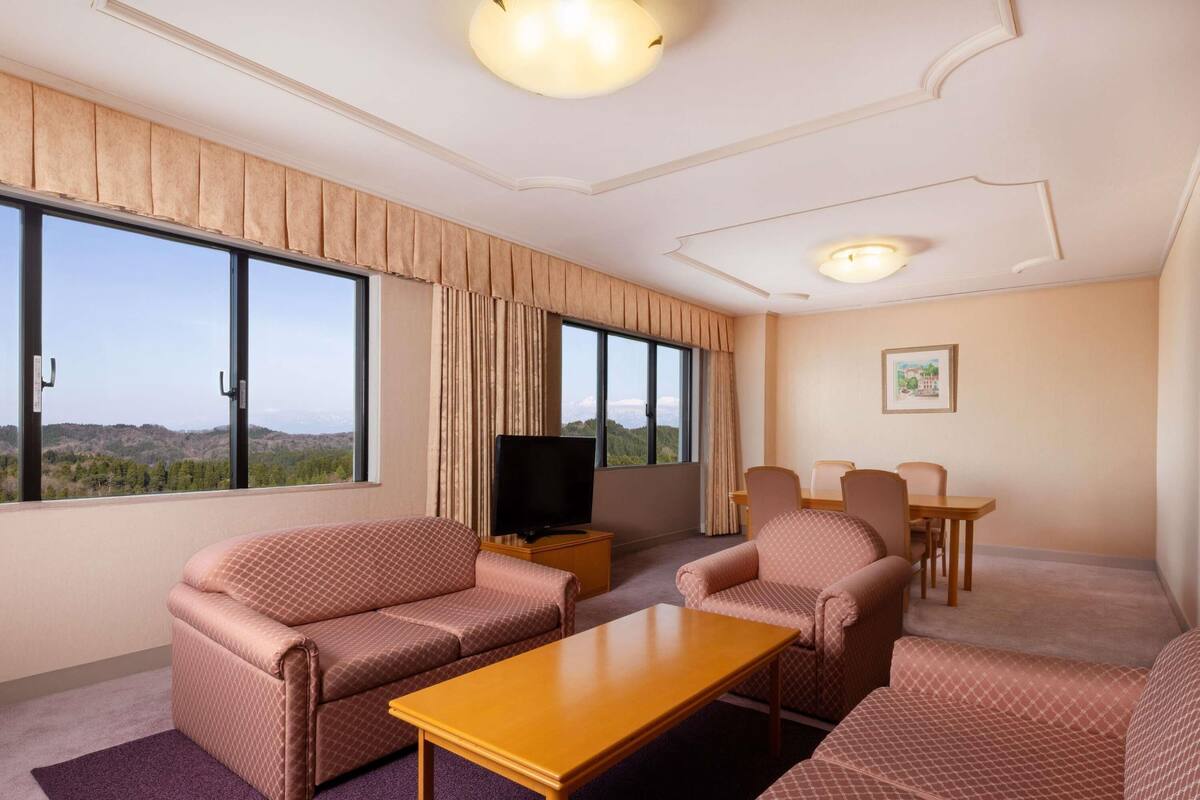Habitación de hotel con un ventanal grande, una televisión de pantalla plana, dos sofás, una mesa baja y una mesa de comedor.