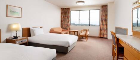 A hotel room with two beds, a desk, a chair, and a view of the outdoors through large windows.