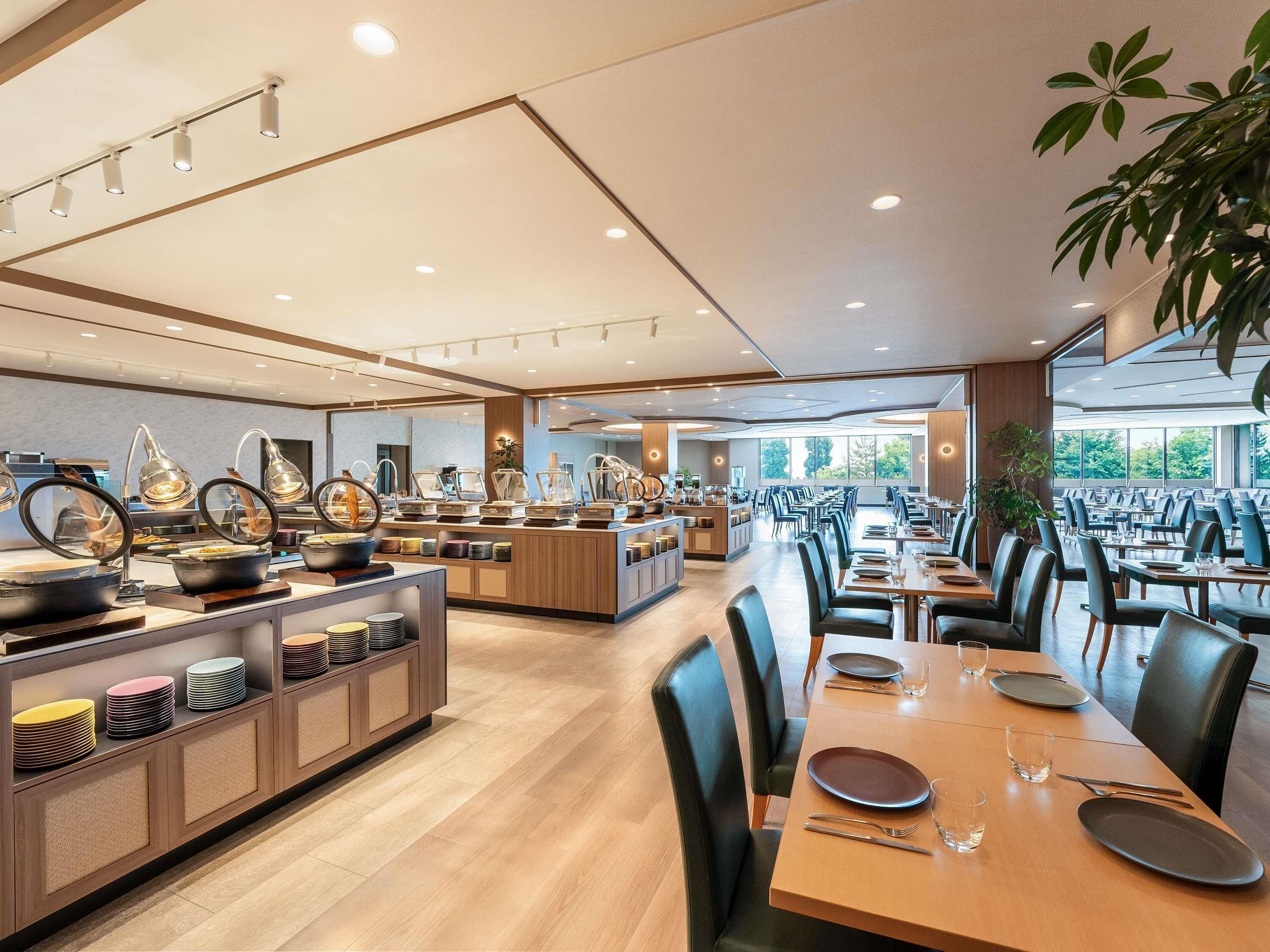 A spacious dining area with multiple tables and chairs, a buffet counter with various food items, and large windows providing natural light.