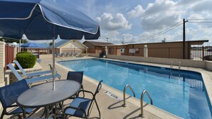 A swimming pool with lounge chairs and umbrellas, a fenced area, and a building in the background. - The Texas Lodge (Gonzales)