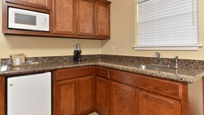 A kitchen with wooden cabinets, a microwave, a coffee maker, and a window with blinds. - The Texas Lodge (Gonzales)