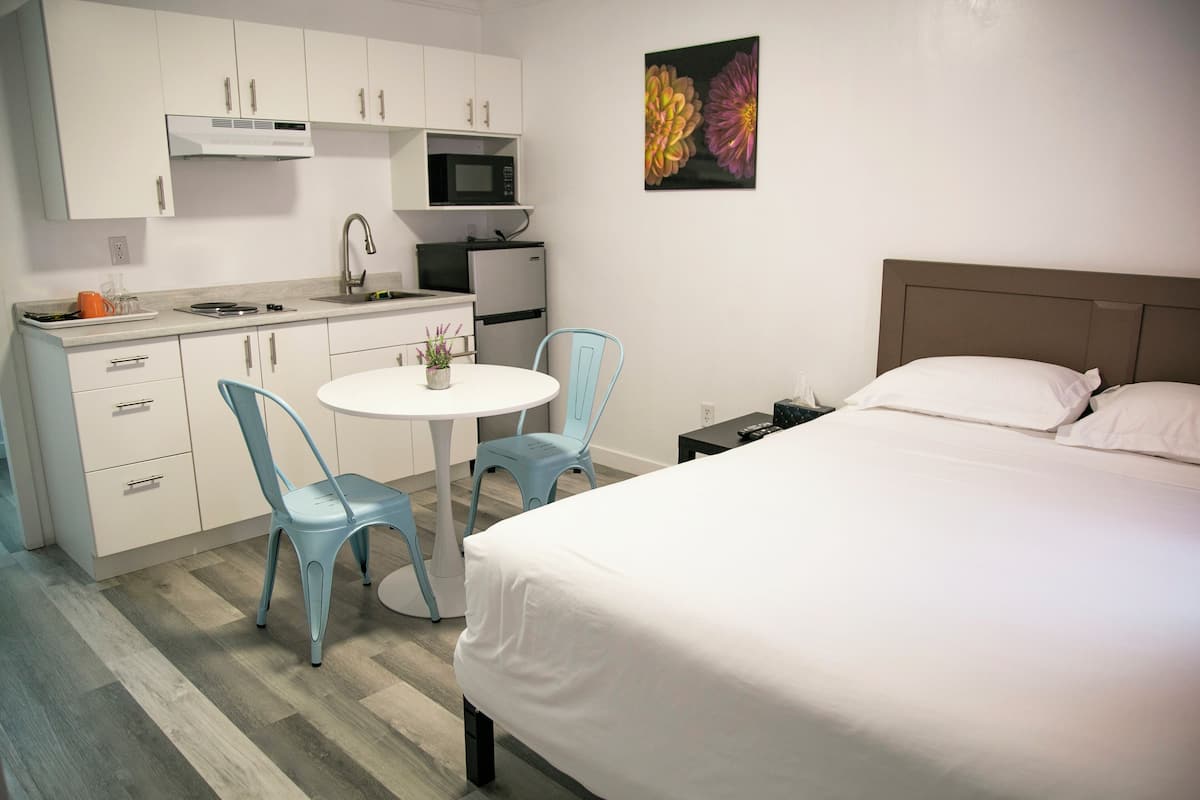 A modern hotel room with a bed, a small kitchenette, a dining table with chairs, and a single artwork on the wall.