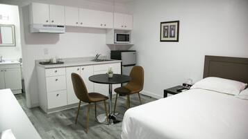 Una camera d'albergo compatta con angolo cottura, un letto, un piccolo tavolo da pranzo con sedie e un comodino con lampada.