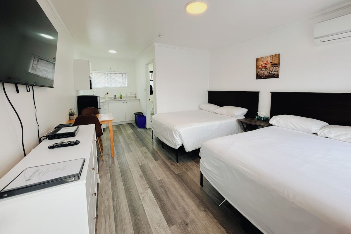 A hotel room with two beds, a desk with a chair, a television mounted on the wall, and a small kitchenette in the background.