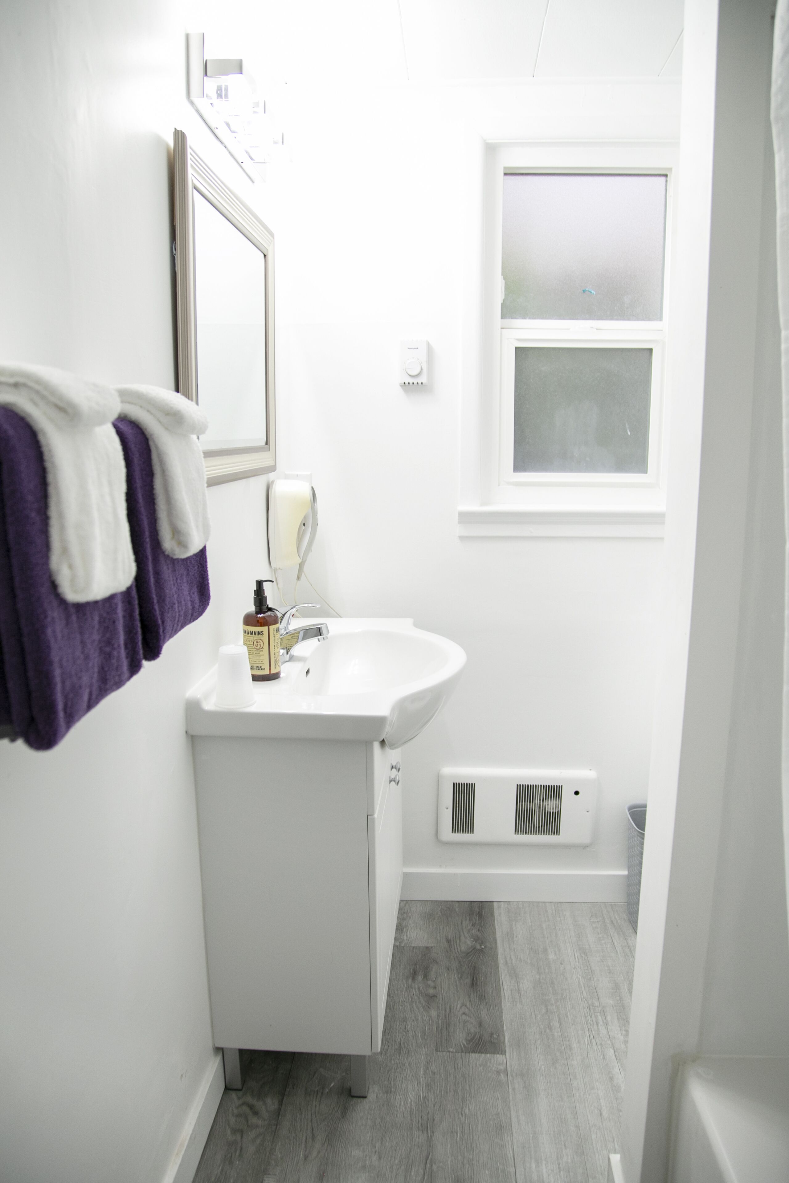 Une petite salle de bain avec un lavabo blanc, un miroir, une fenêtre et un porte-serviettes avec des serviettes violettes.