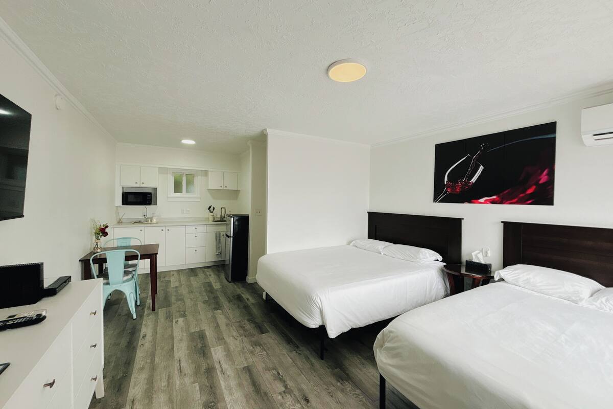 A hotel room with two beds, a kitchenette, a TV, and a painting on the wall.