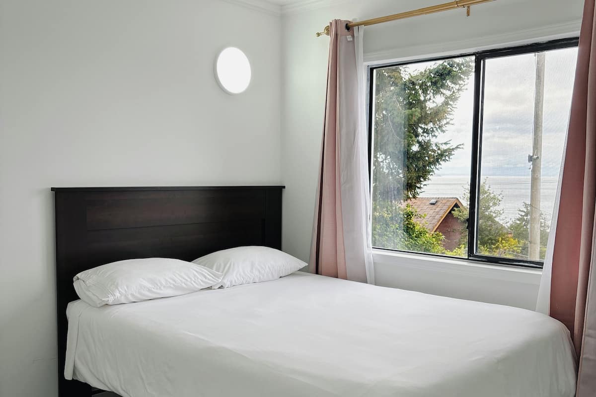 A neatly made bed with white bedding in a room with a dark wooden headboard, a round wall light, and a window with curtains.