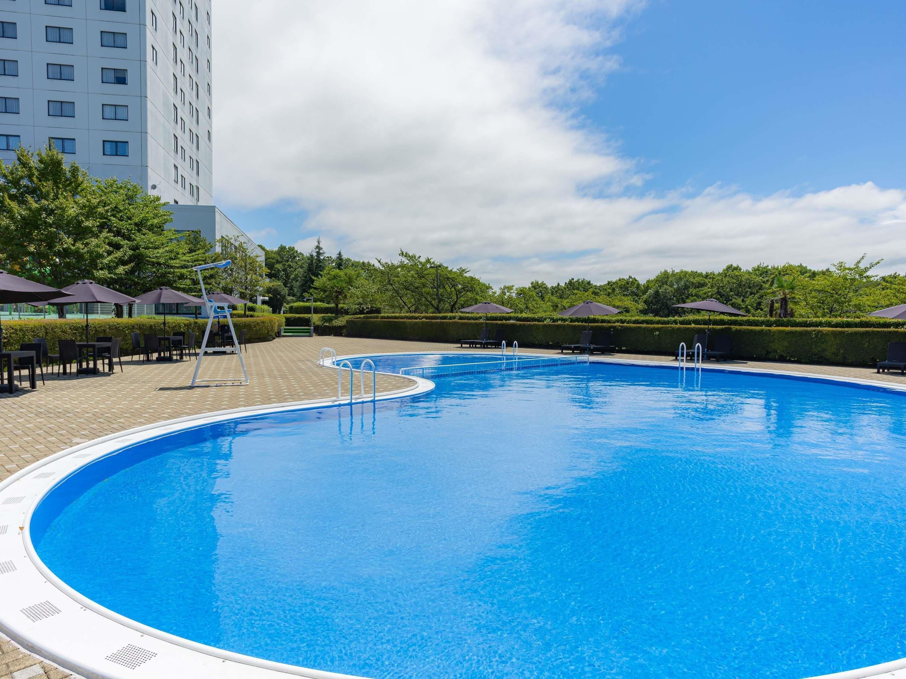 A swimming pool with a clear blue water surface, surrounded by lounge chairs and umbrellas, with a building in the background.