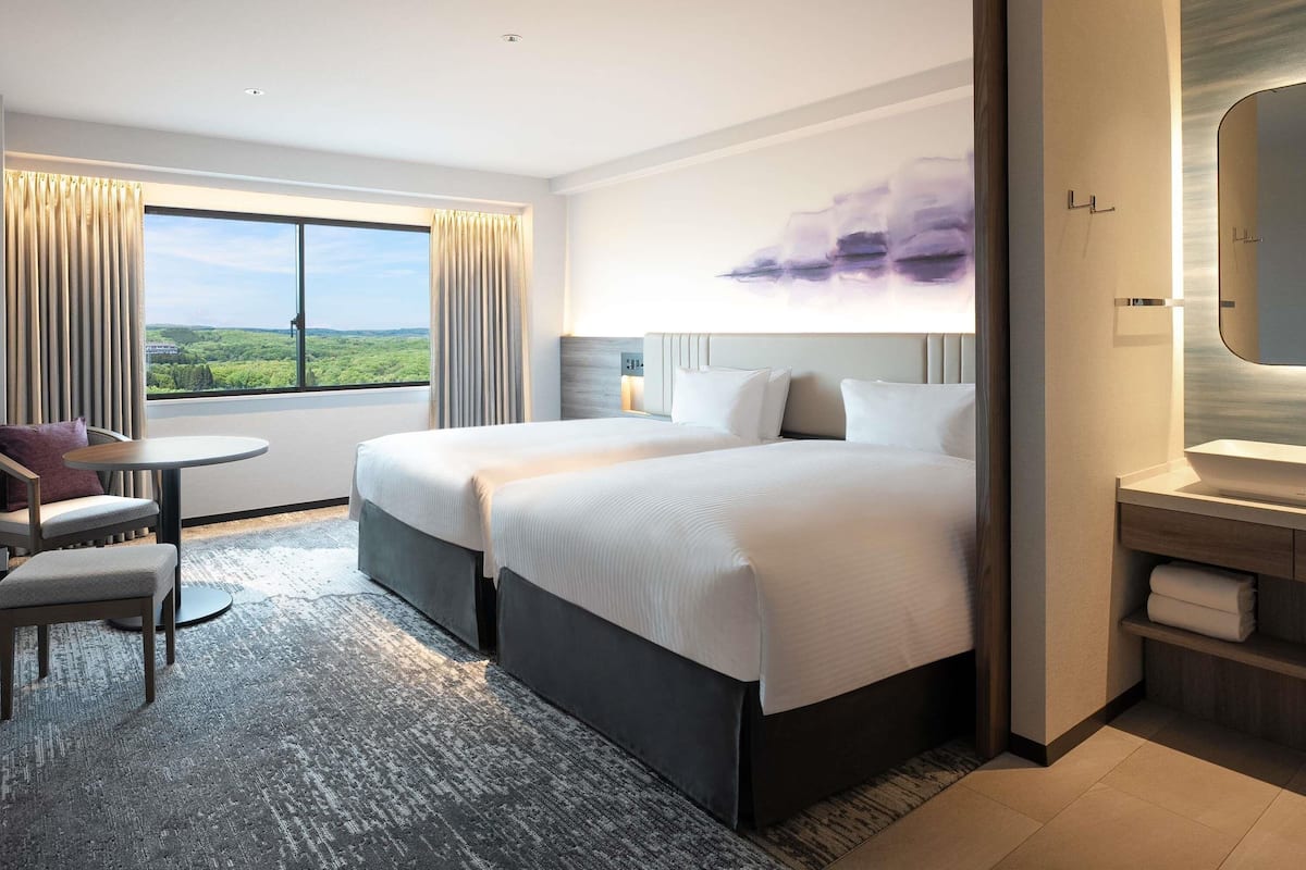 A hotel room with two beds, a sitting area with a table, a large window with a view, and a bathroom with a mirror and sink.