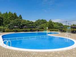 Ein großer Außenpool mit klarem, blauem Wasser, umgeben von Liegestühlen und Sonnenschirmen, mit einer Hecke im Hintergrund.