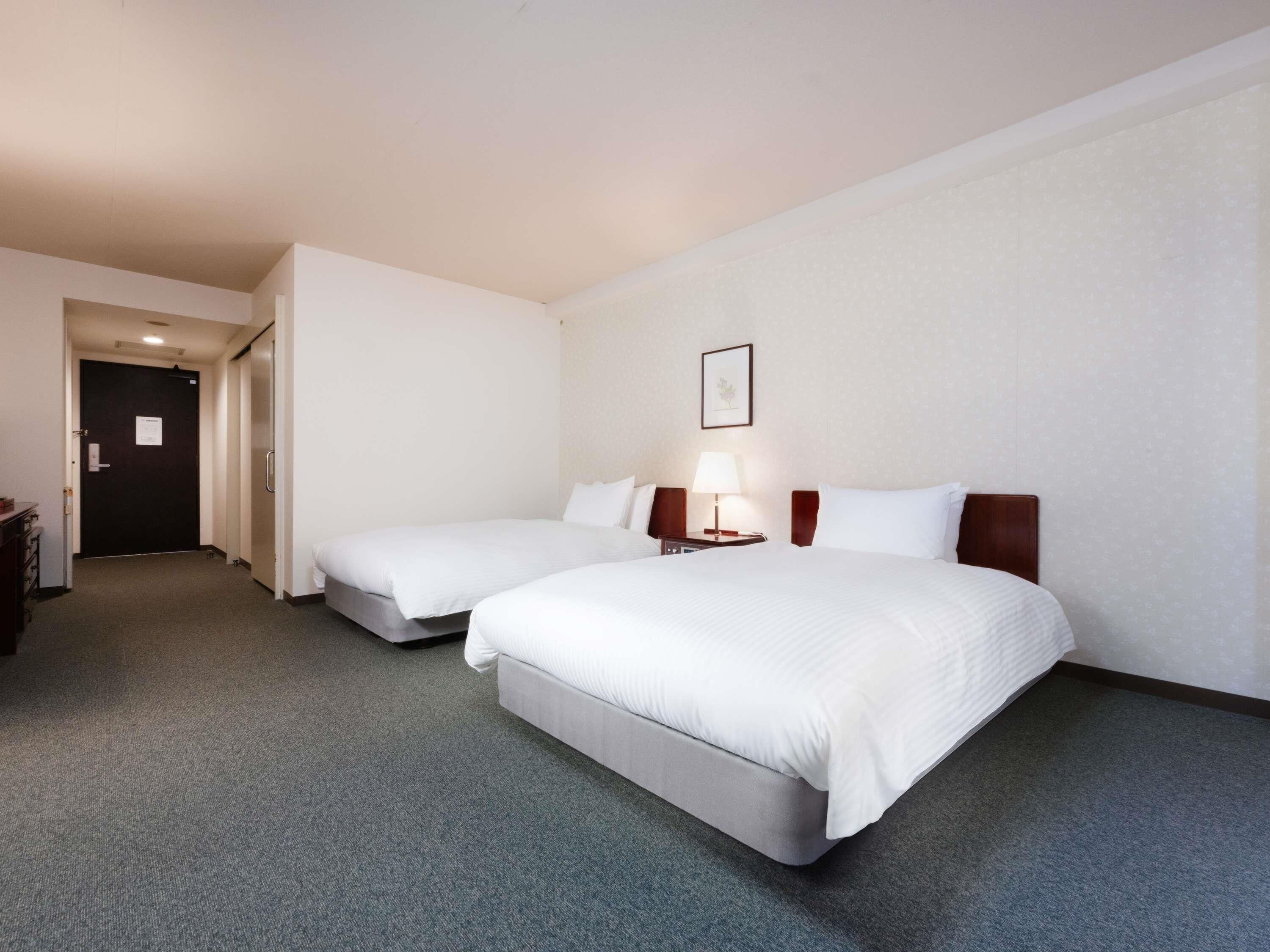 A hotel room with two beds, a desk, a chair, a lamp, and a door.