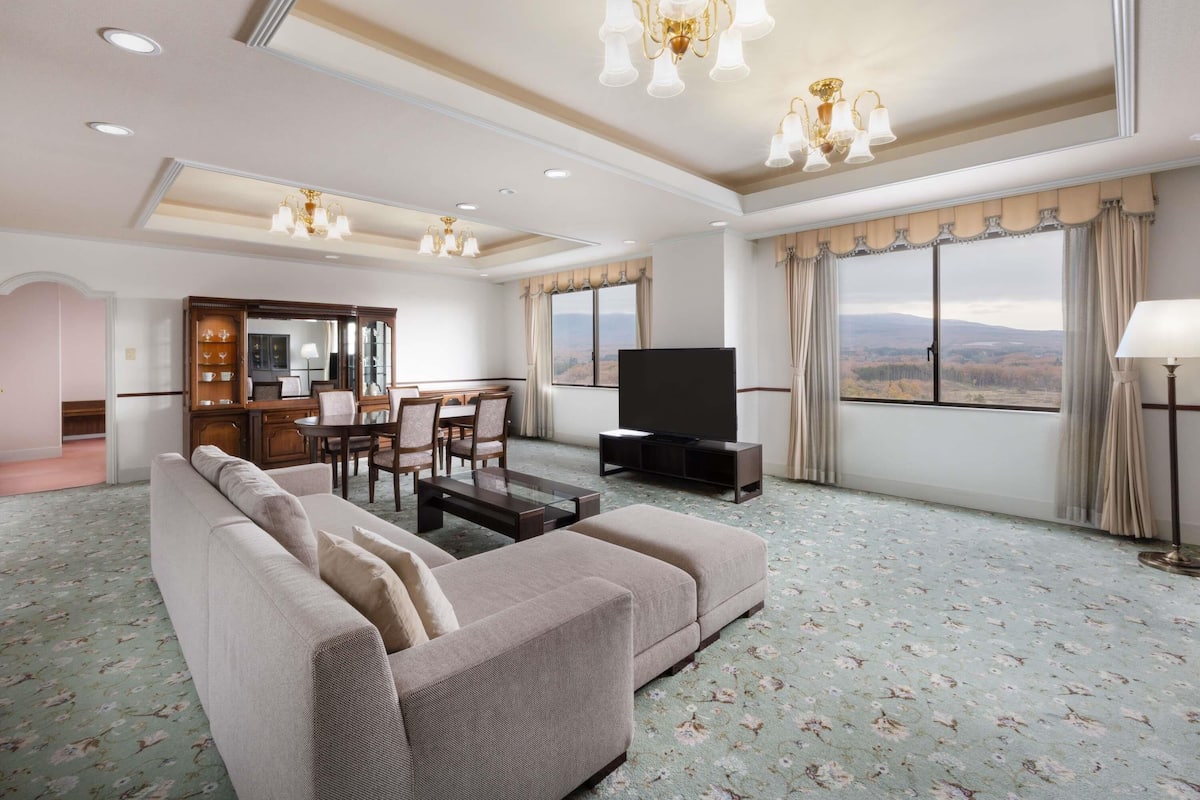 A spacious living room with a sofa, a flat-screen TV, a dining area with a table and chairs, and a view of the mountains through large windows.