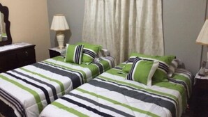 A hotel room with two beds, each with striped bedding in green, white, and black. There is a window with curtains, a bedside table with a lamp, and a dresser. - Sunset Oasis (Derricks)