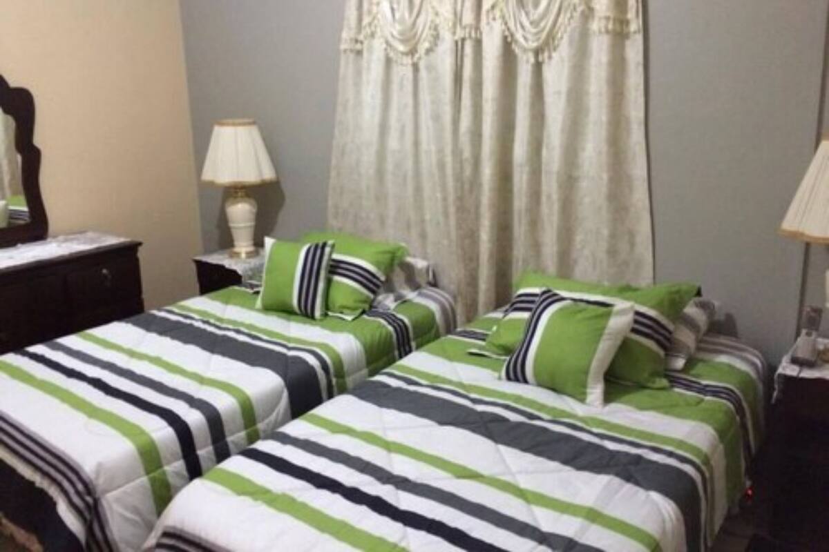 Habitación de hotel con dos camas, cada una con ropa de cama a rayas en verde, blanco y negro. Hay una ventana con cortinas, una mesita de noche con lámpara y un aparador.