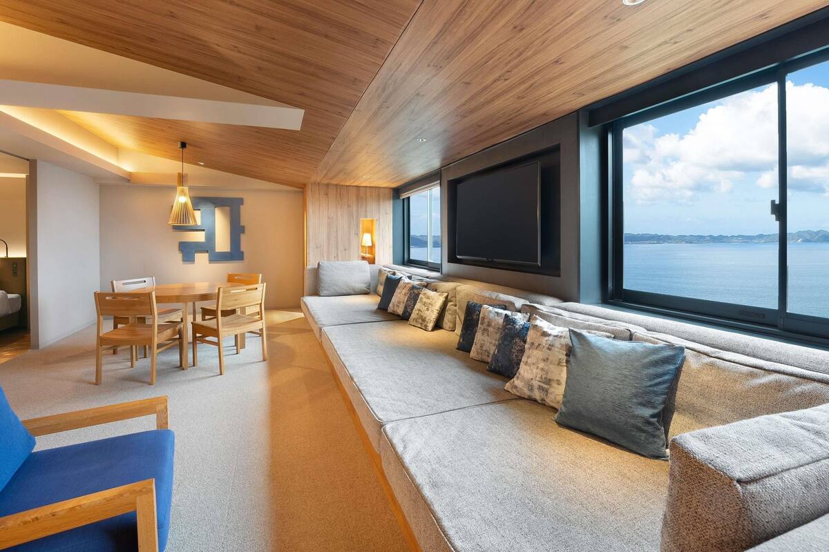 A modern living room with a wooden ceiling, a large window with a view of the sea, a sofa with cushions, and a dining area with chairs and a table.