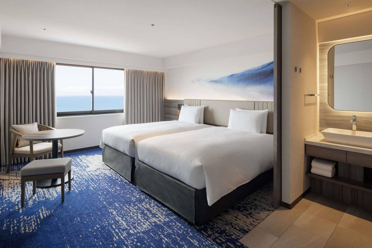 A hotel room with two beds, a small table, a chair, a bathroom with a sink, and a view of the sea.