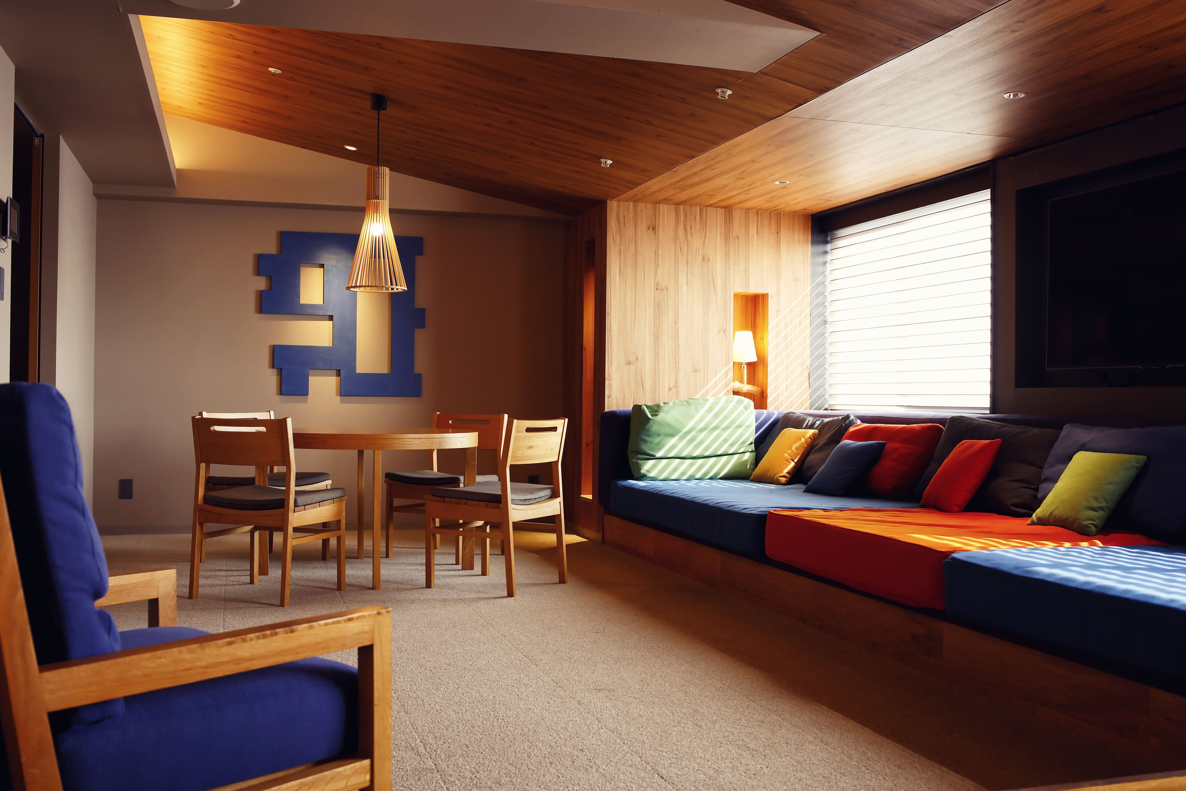 A modern living room with a wooden ceiling, a blue sofa with colorful cushions, a dining table with chairs, and a wall-mounted lamp.