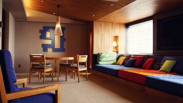 A modern living room with a wooden ceiling, a blue sofa with colorful cushions, a dining table with chairs, and a wall-mounted lamp.