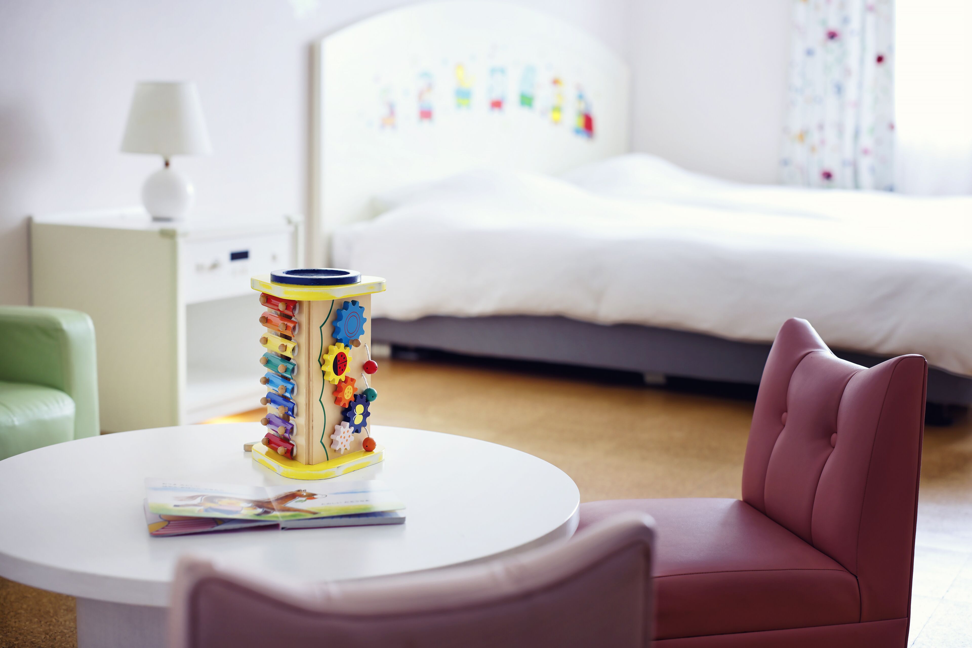 Una habitación de hotel con cama, mesita de noche, lámpara, una mesa redonda con una revista y un juguete colorido.