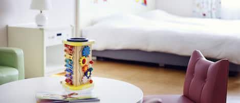 Una habitación de hotel con cama, mesita de noche, lámpara, una mesa redonda con una revista y un juguete colorido.