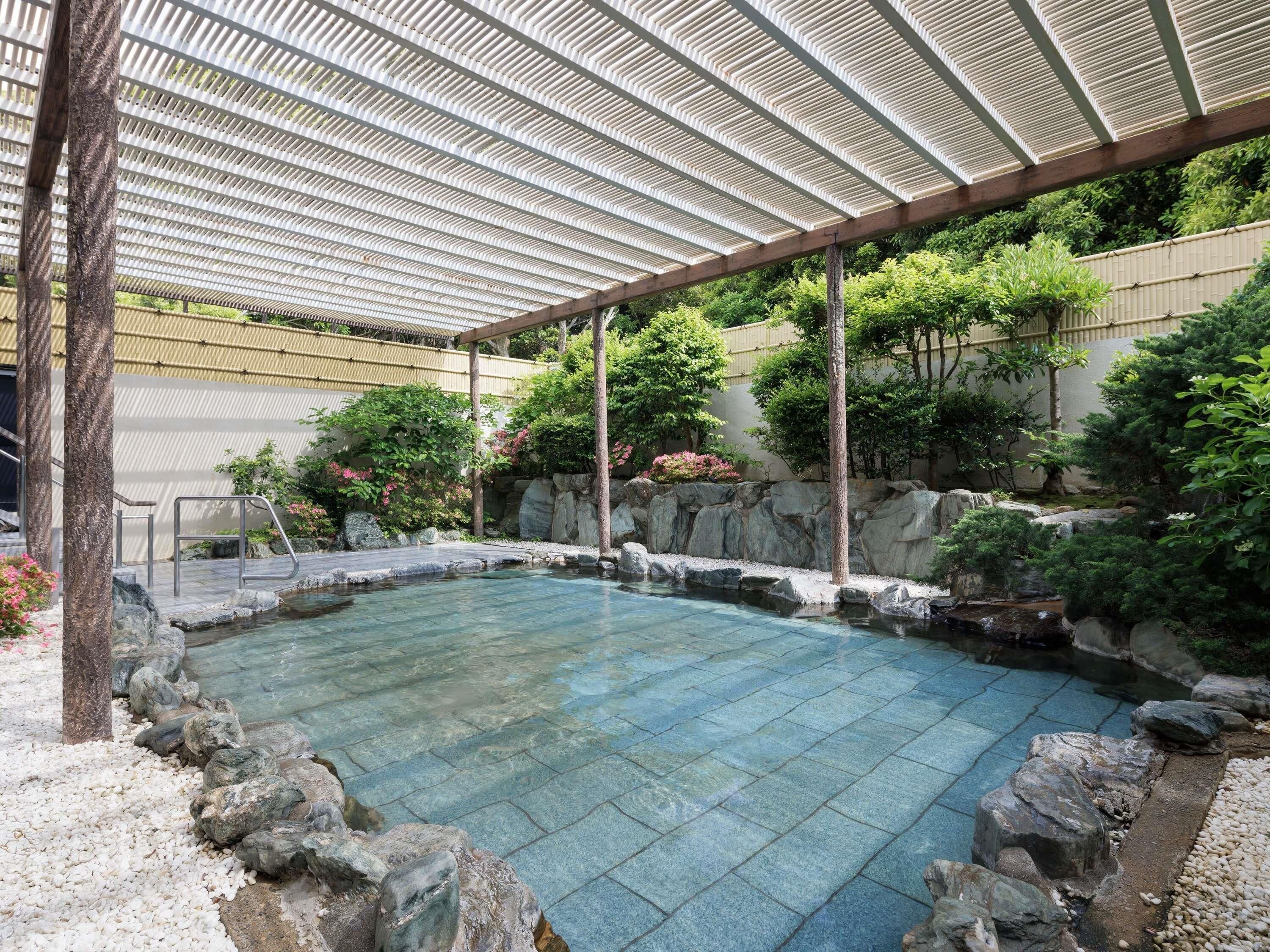 A clear pool with a stone border, surrounded by rocks and greenery, under a covered area with a metal roof.