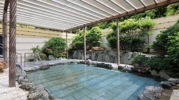 A clear pool with a stone border, surrounded by rocks and greenery, under a covered area with a metal roof.