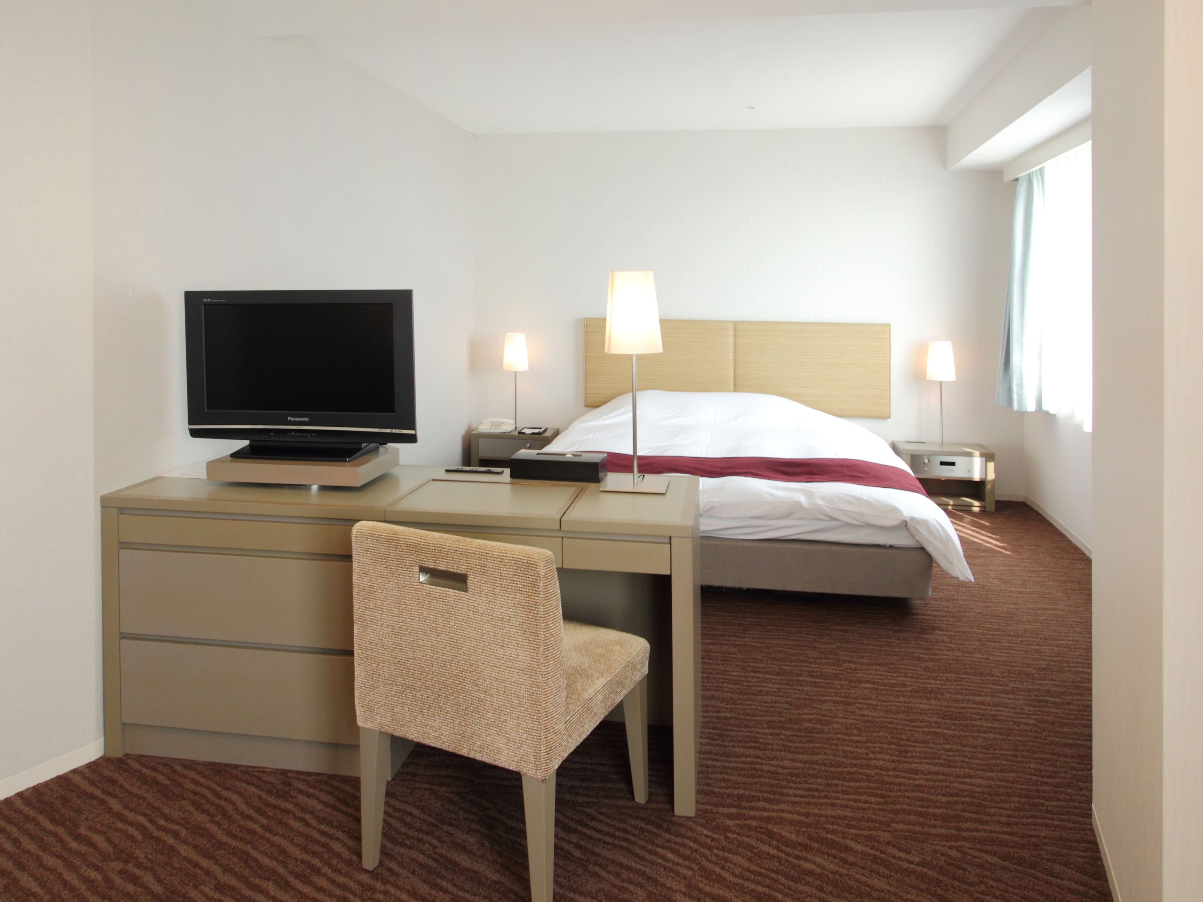 A hotel room with a bed, a desk with a television, a chair, and two bedside lamps.