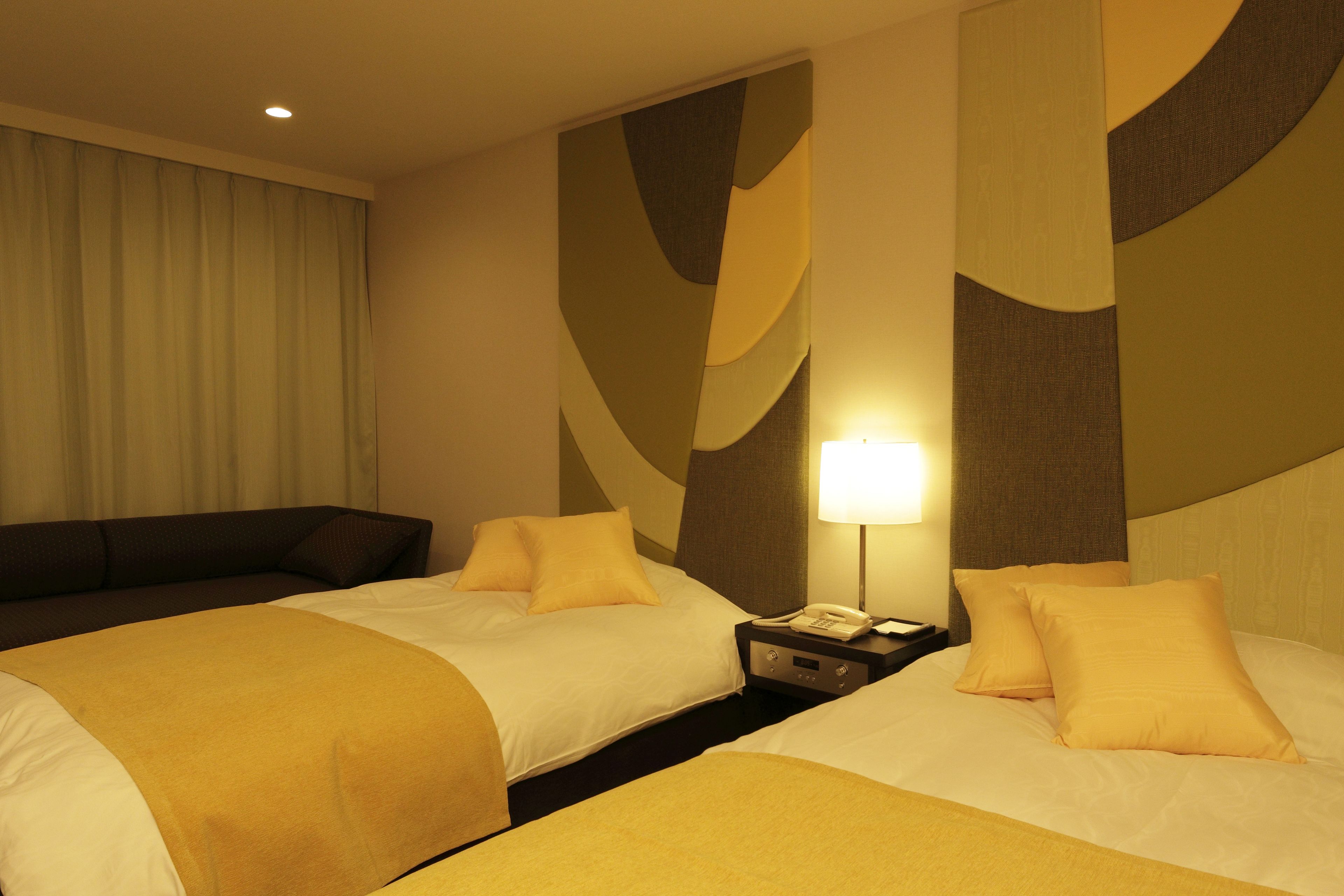 A hotel room with two beds, a nightstand, a lamp, and a large window with curtains.