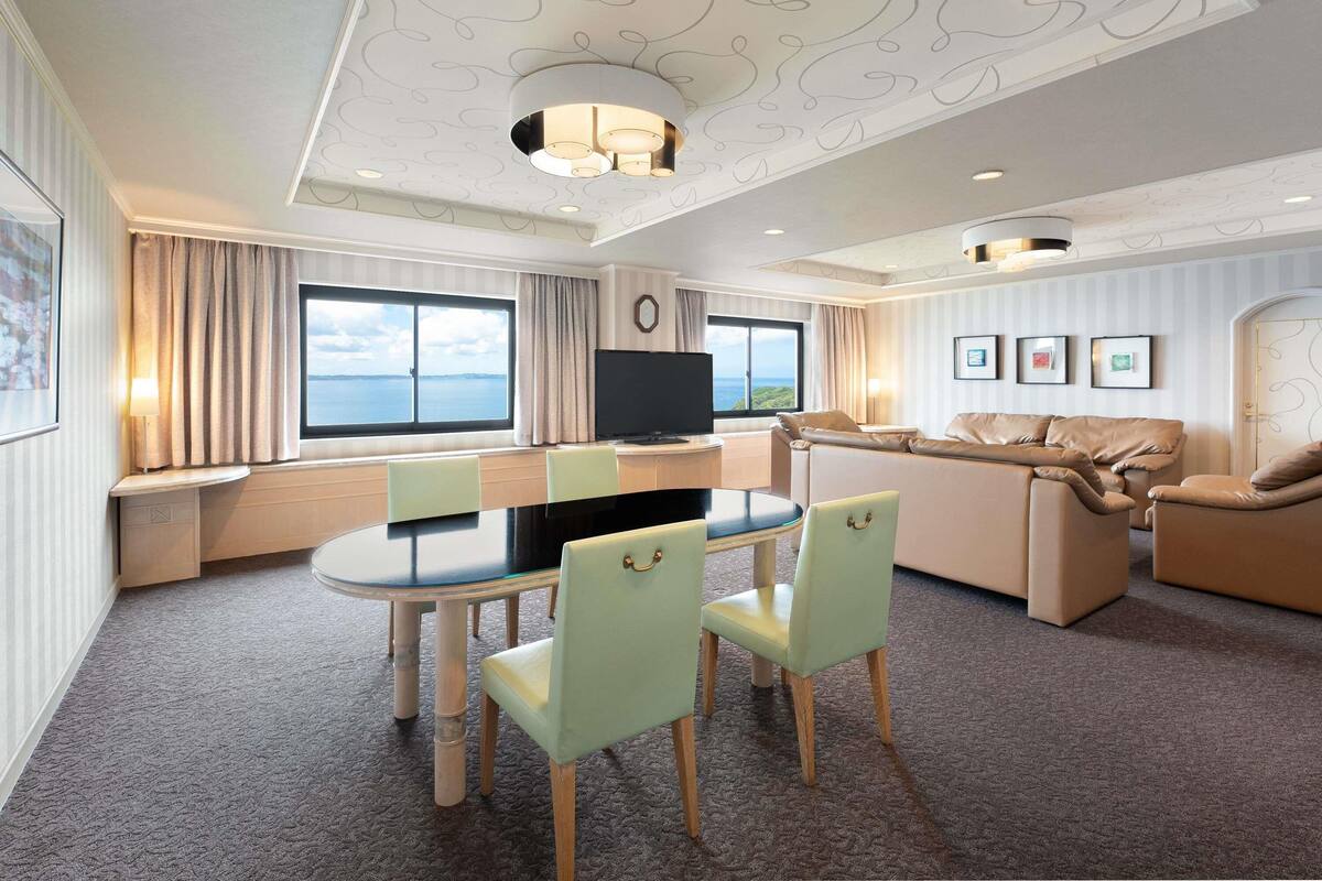 A modern hotel room with a dining area, a sofa set, a TV, and large windows offering a view of the sea.