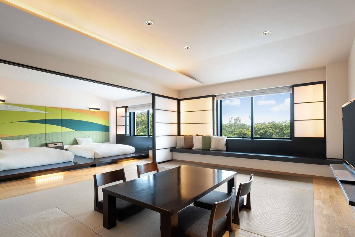 A modern hotel room with a large window, a sofa, a dining table, and a decorative wall.