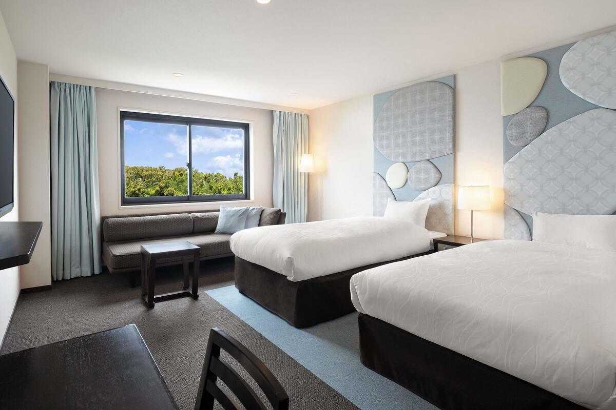A hotel room with two beds, a sofa, a desk, and a large window offering a view of greenery.