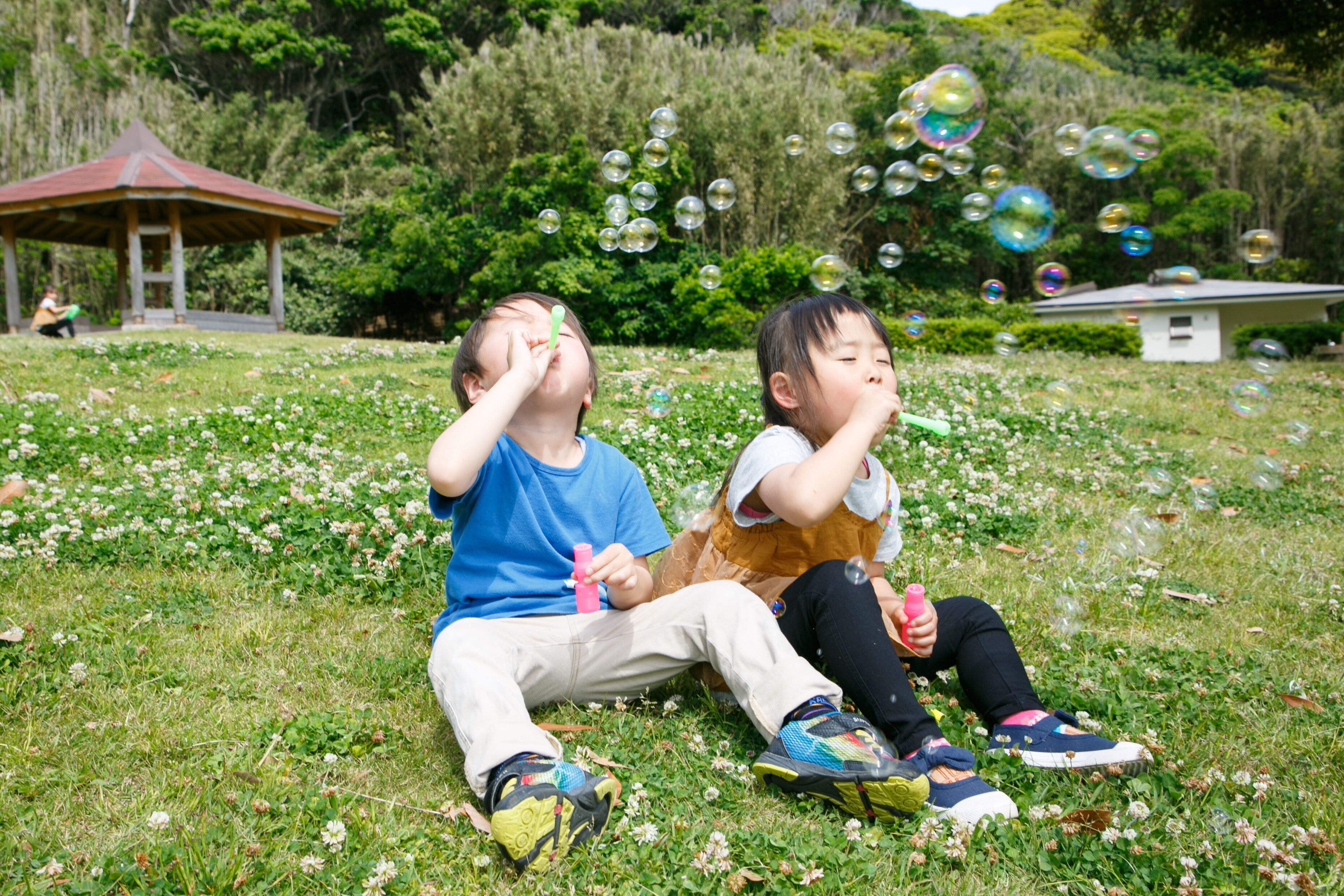 Dos niños jugando con burbujas en un césped, con un pabellón al fondo.