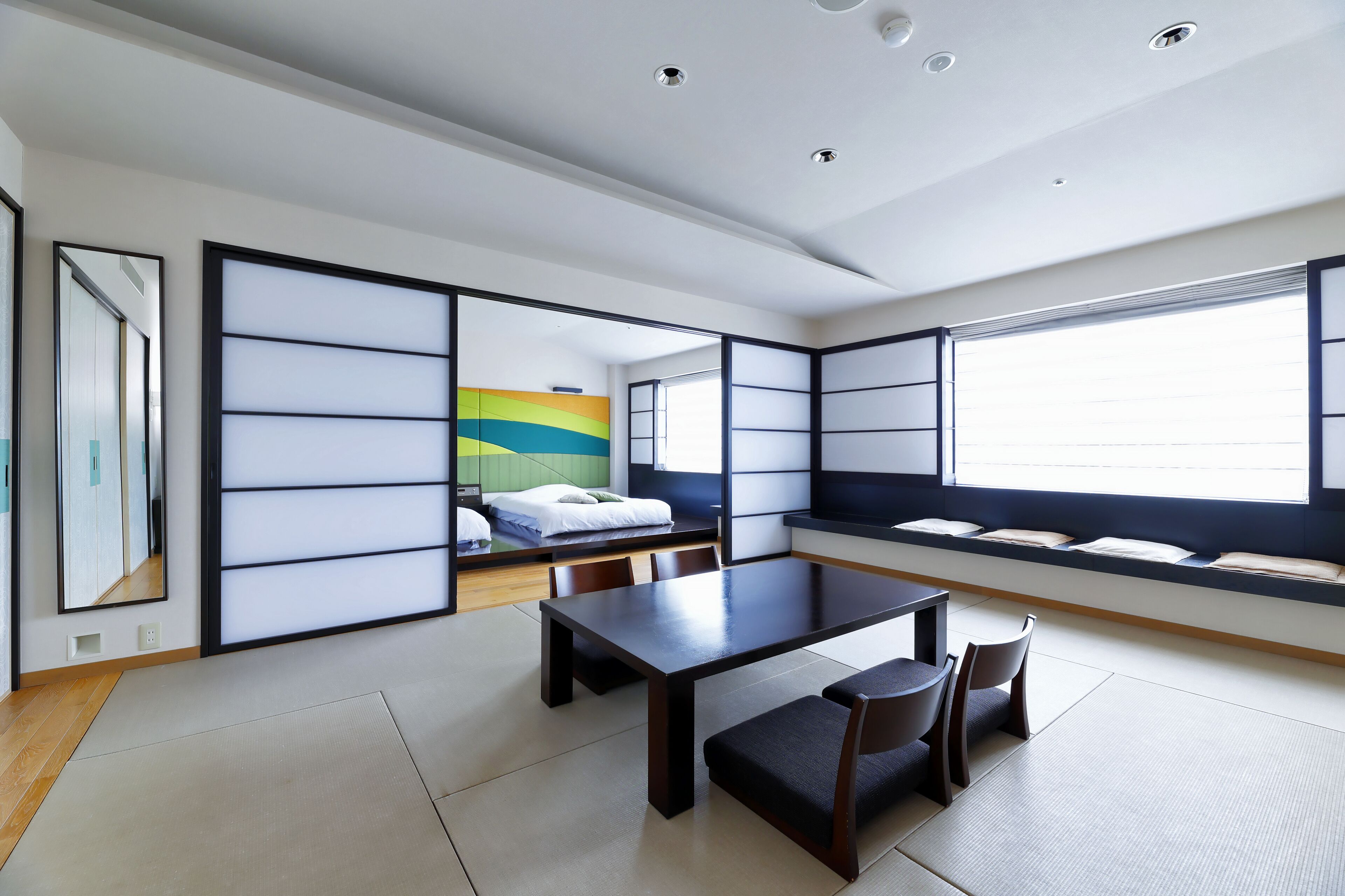 A modern room with tatami flooring, a low table with chairs, a bed with a colorful headboard, and large windows with blinds.