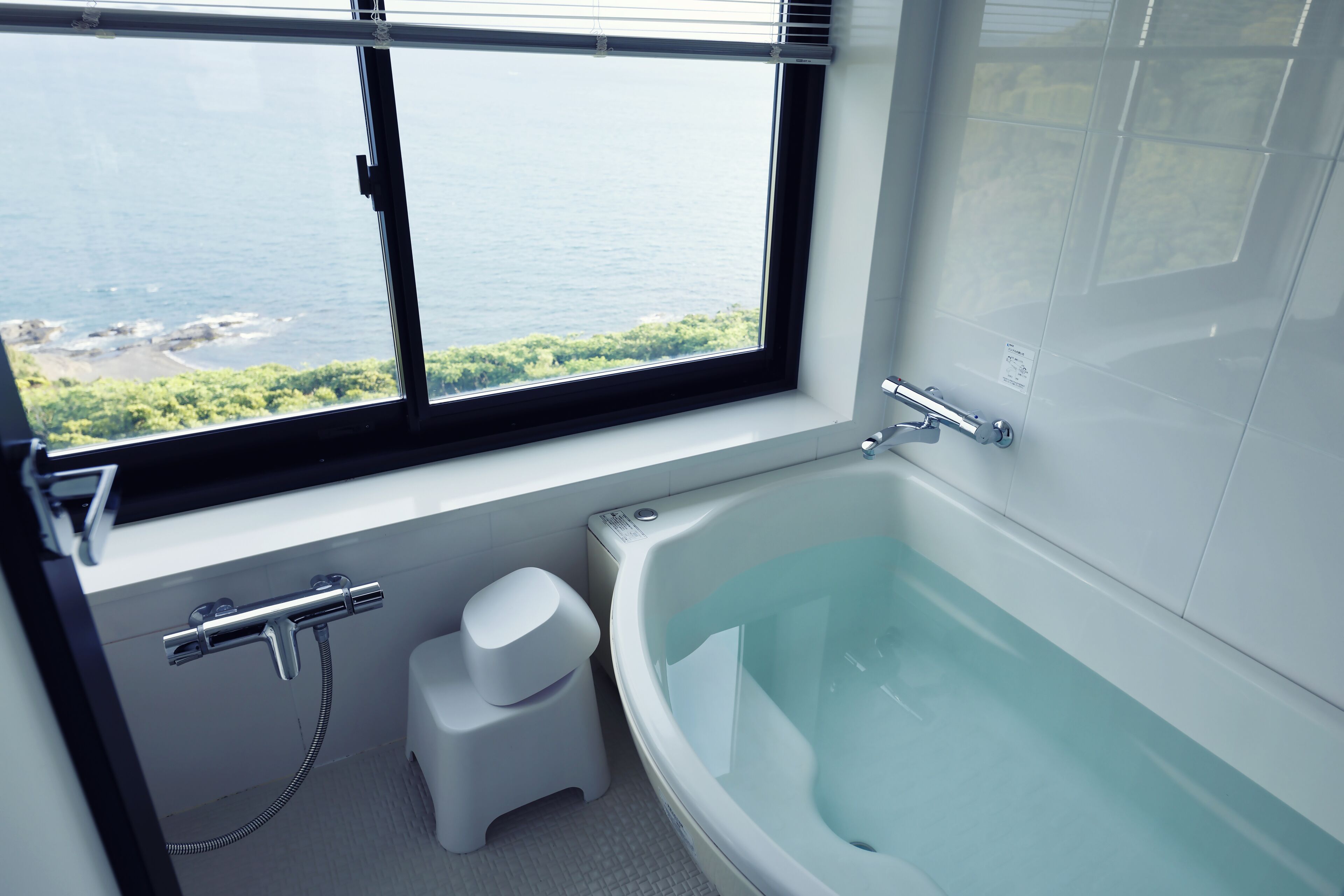 Ein modernes Badezimmer mit einer großen Badewanne, einer Dusche und einer Toilette. Ein Fenster bietet Blick auf das Meer und die grüne Landschaft.