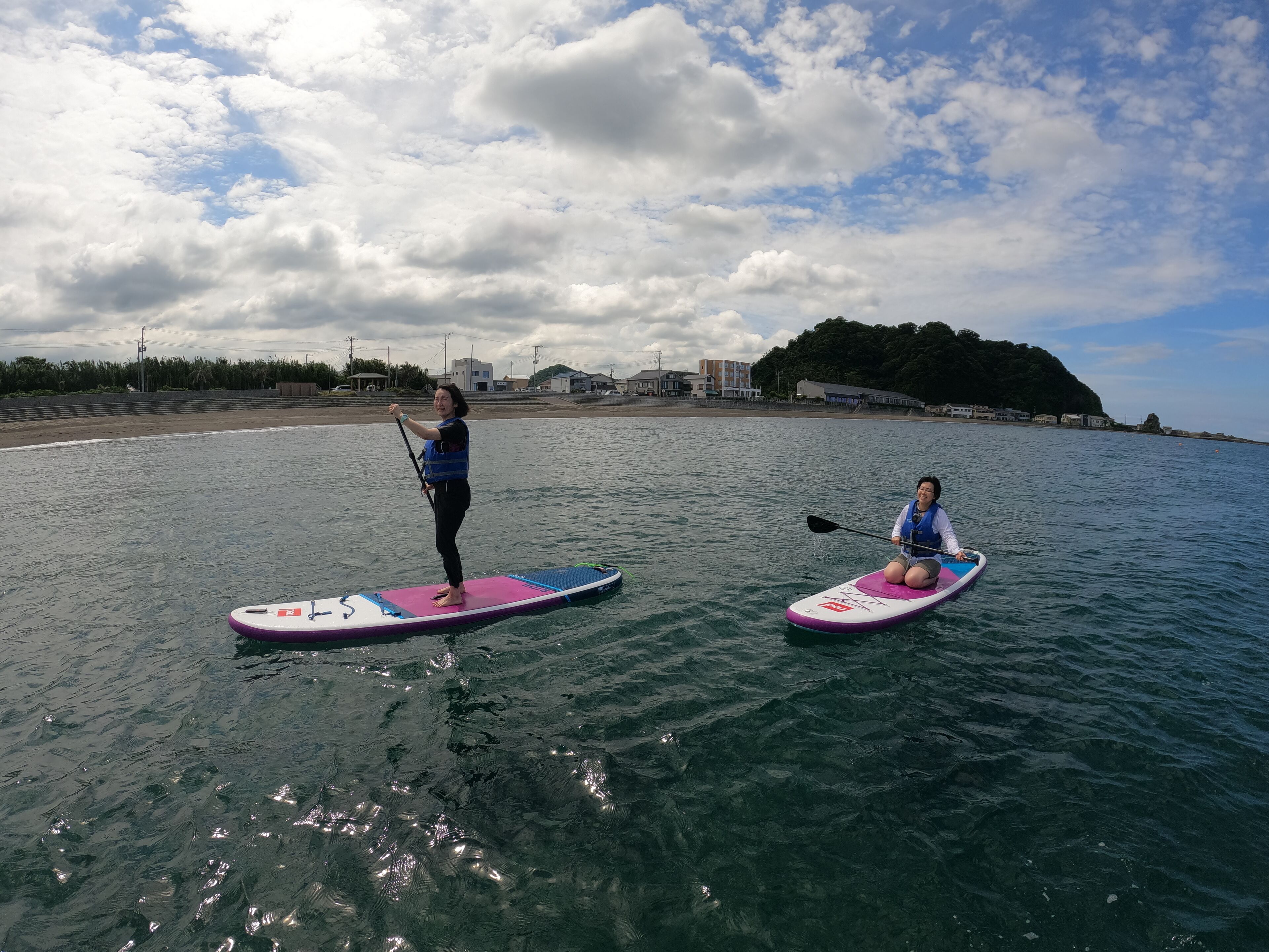 Deux personnes pratiquent le paddle sur une étendue d’eau calme, avec une plage et des bâtiments en arrière-plan.