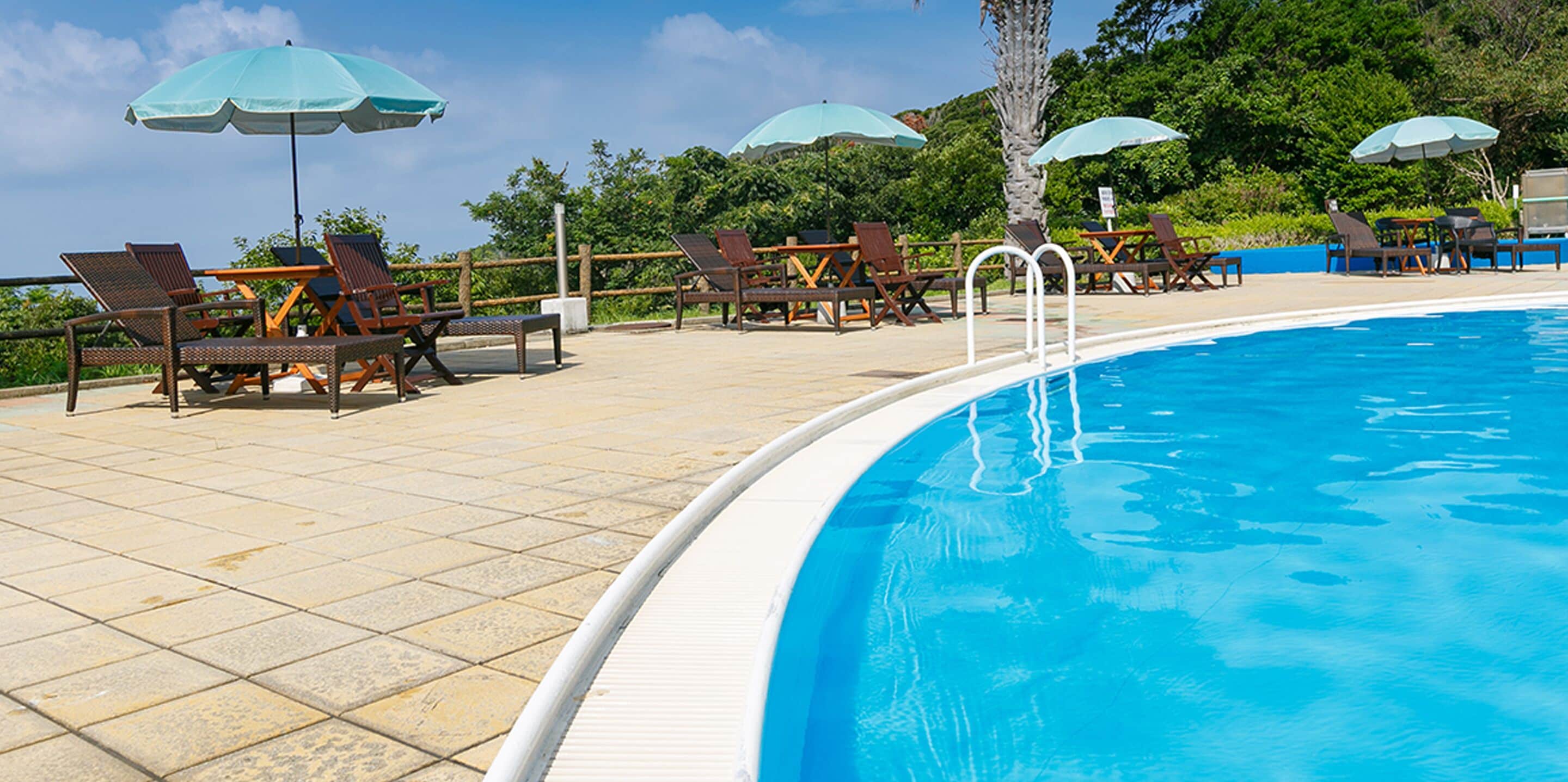 Piscina con sillones de descanso y sombrillas, rodeada de una terraza de baldosas y vegetación.