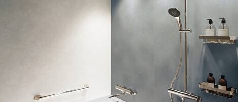 Une salle de bain moderne avec une baignoire blanche, une pomme de douche et un sèche-mains. On peut voir deux bouteilles sur une étagère.