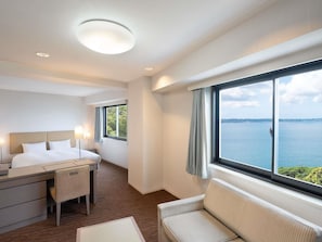 A hotel room with a bed, desk, chair, sofa, and a large window with a view of the sea.