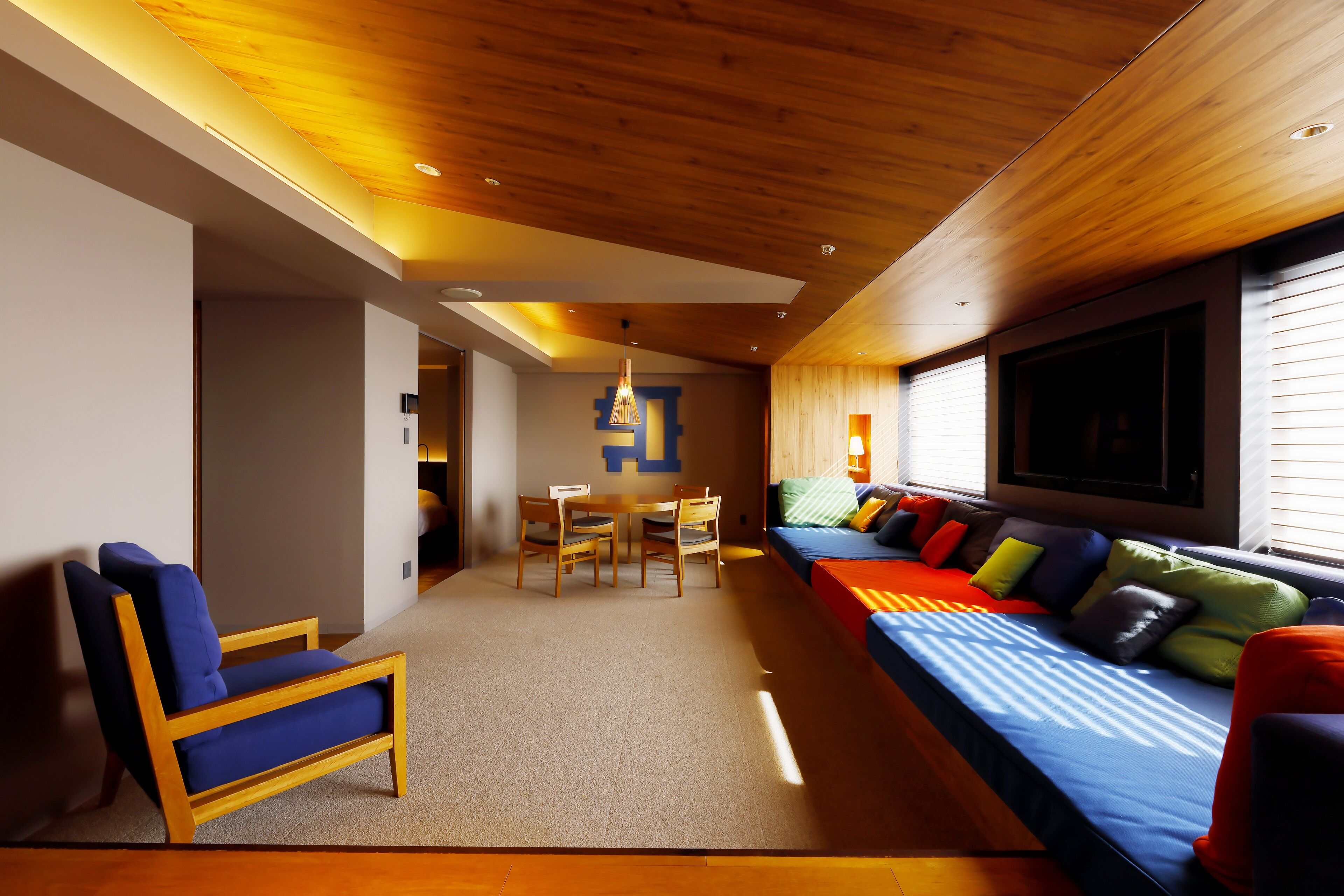 A modern living room with a wooden ceiling, a sofa with colorful cushions, a wooden chair, and a table with chairs.