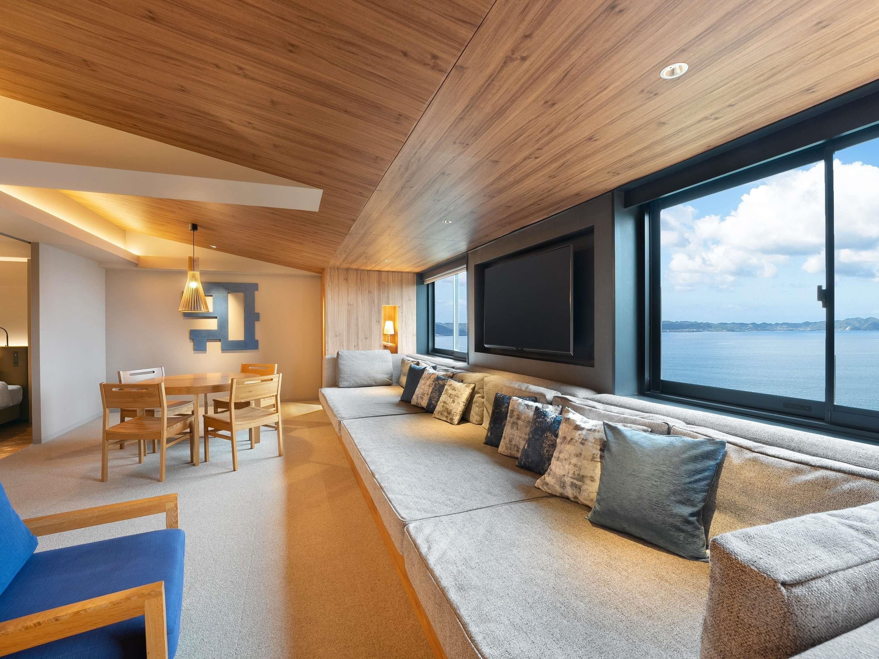 Un soggiorno moderno con soffitto in legno, un'ampia finestra vista mare, un divano con cuscini e un'area pranzo con sedie e tavolo.