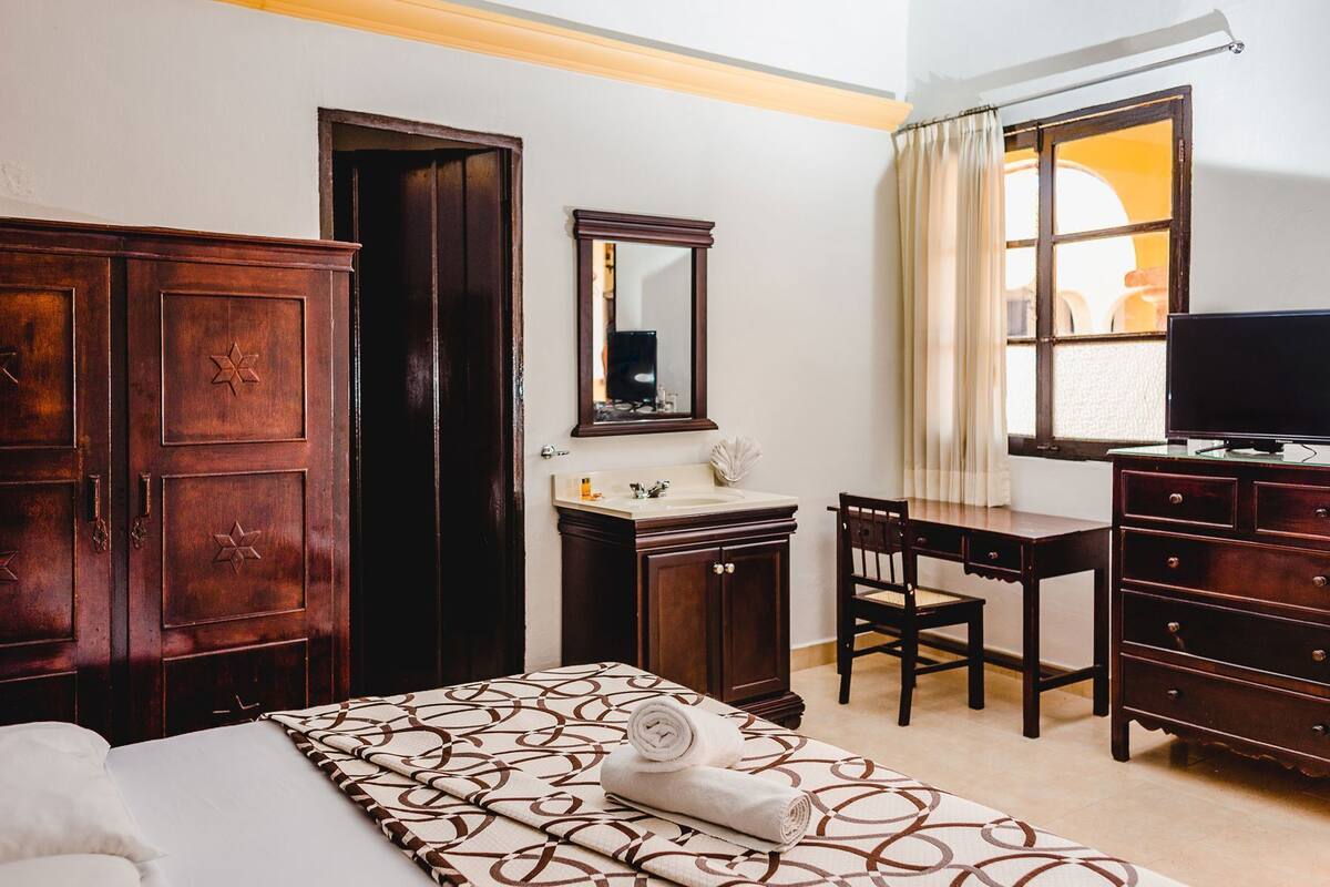 A hotel room with a bed, a desk, a dresser, a mirror, a television, and a window with curtains.