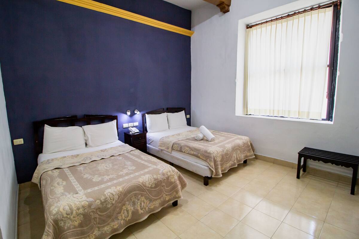 A room with two beds, a bench, a window with curtains, and a nightstand with a lamp.